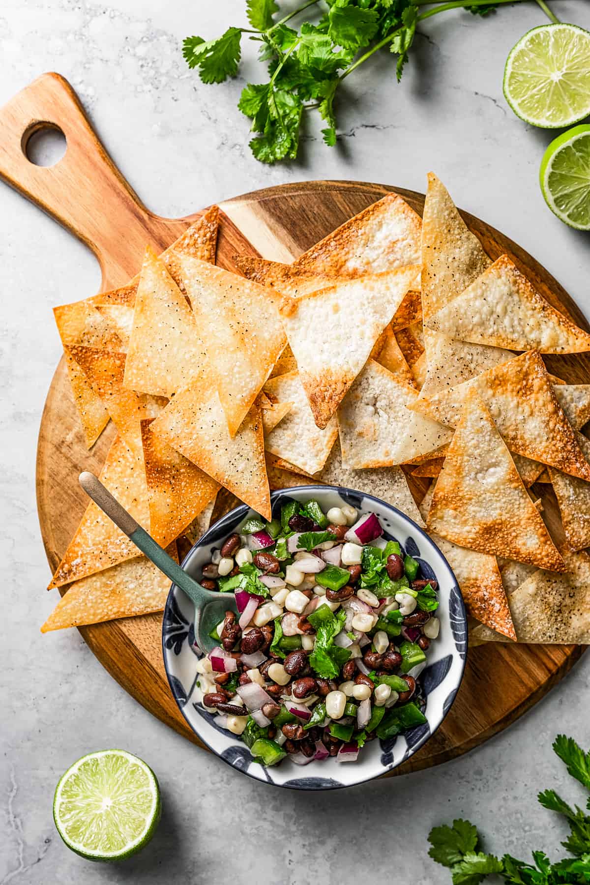 Overhead view of a wooden tray covered in wonton chips, with a bowl of corn salsa, surrounded by limes and cilantro.