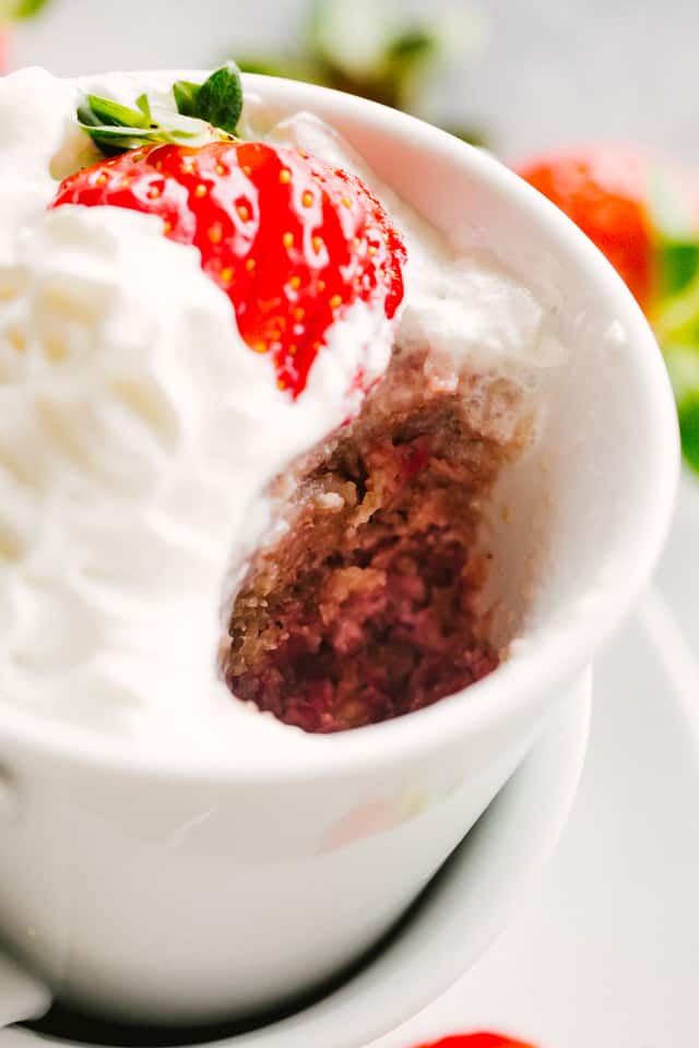 A white mug with cake baked inside it, topped with whipped cream and a sliced strawberry. A scoop of cake has been taken out, showing the texture of the inside of the cake.