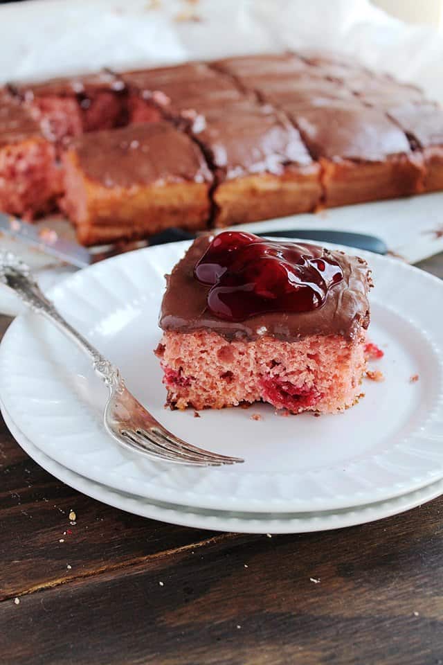 A single slice of cherry cake on a plate with chocolate frosting and a gooey cherry on top