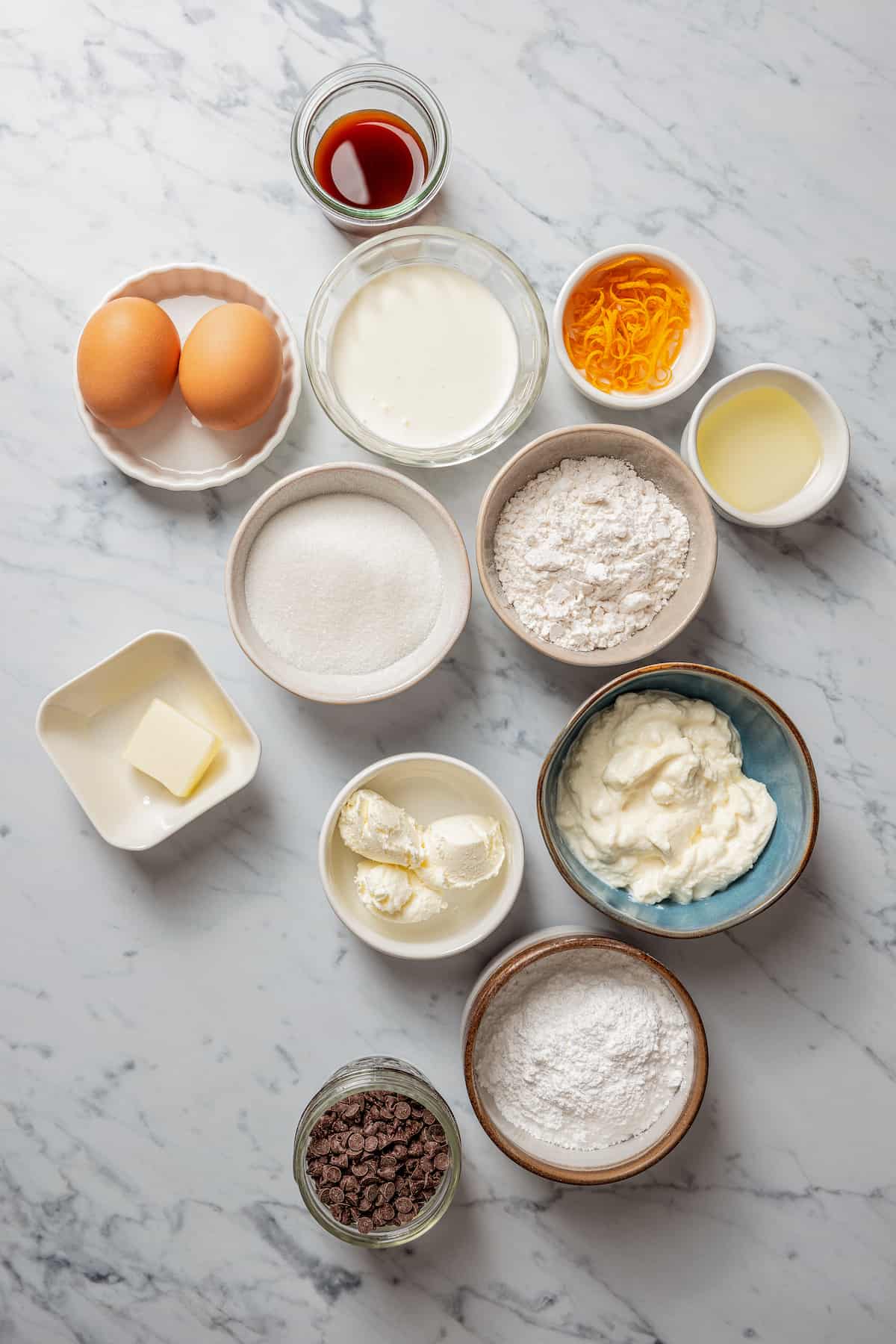 Overhead view of the ingredients needed to make cannolis: a bowl of vanilla extract, a bowl of orange zest, a bowl of eggs, a bowl of heavy cream, a bowl of sugar, a bowl of powdered sugar, a bowl of ricotta, a bowl of cream cheese, a bowl of butter, a bowl of flour, and a bowl of chocolate chips