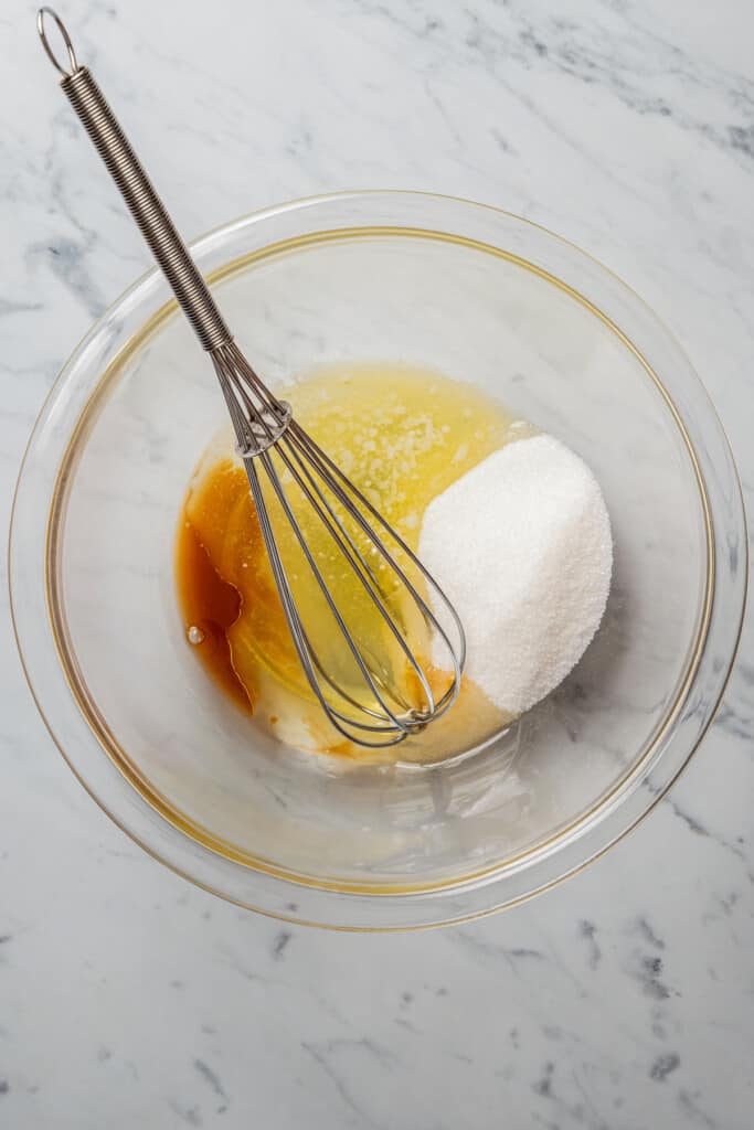 A mixing bowl with sugar, egg whites, melted butter, vanilla, and a whisk
