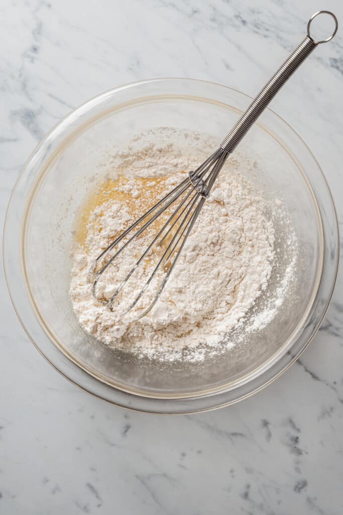 A mixing bowl with a pile of flour on top of wet ingredients, with a whisk