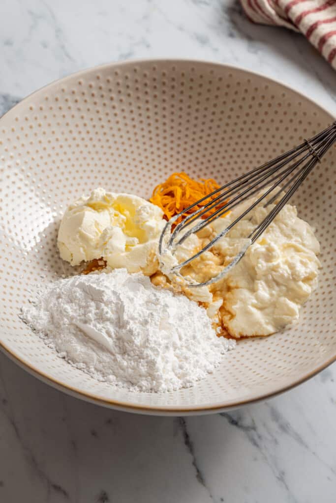 A bowl with ricotta, orange zest, cream cheese, powdered sugar, and a whisk