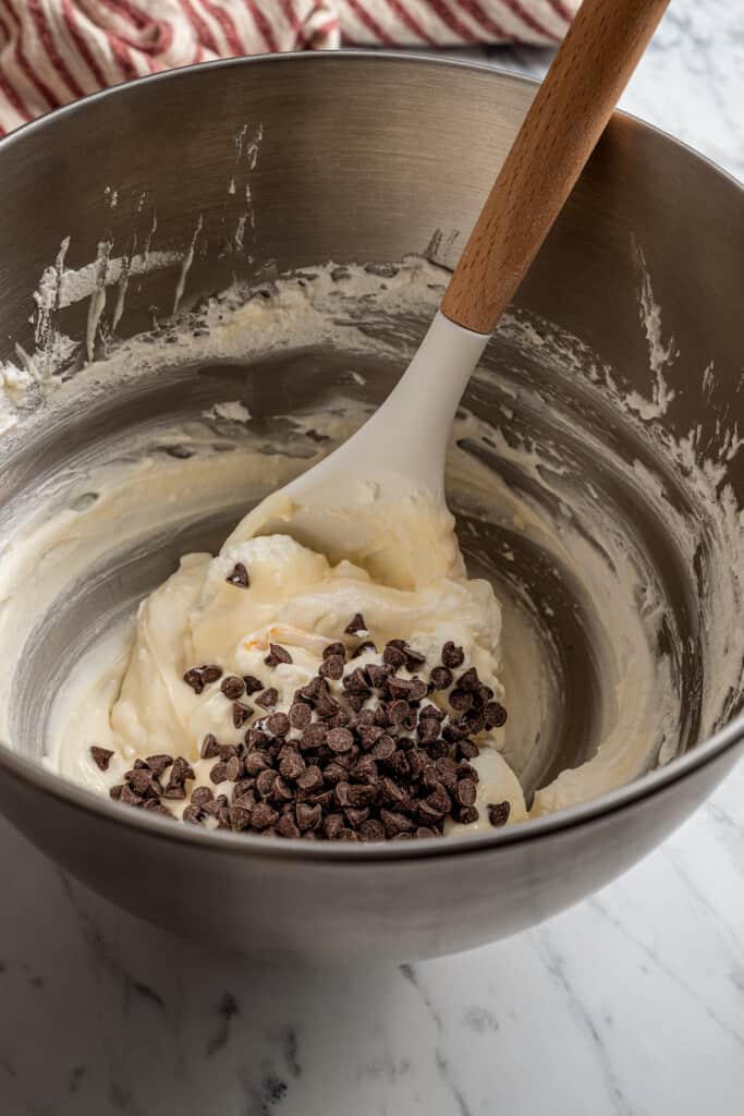 A mixing bowl with chocolate chips and whipped cream being folded into cannoli filling with a spatula