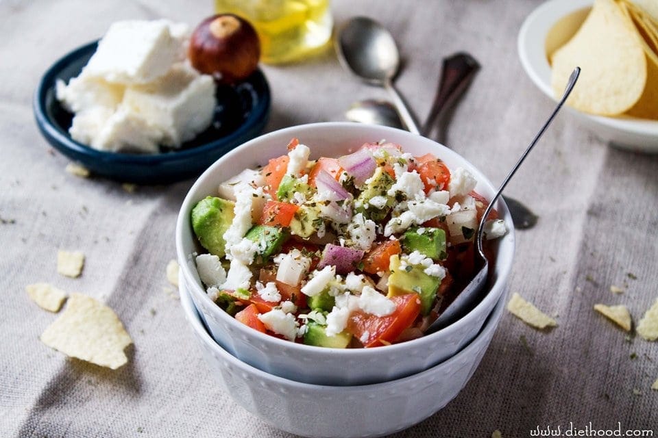 A ramekin filled with fresh avocado salsa in front of a dish filled with chips and another filled with feta cheese.