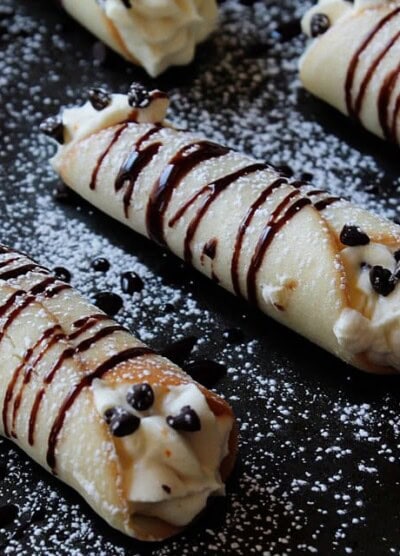 A row of cannolis drizzled with chocolate and dusted with powdered sugar