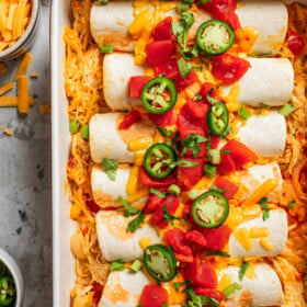 Enchiladas in a baking dish and topped with tomatoes, cheese, and sliced jalapenos.
