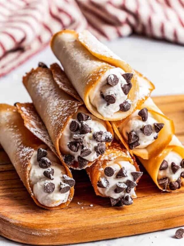 Six cannolis forming a pyramid on a cutting board with a kitchen towel in the background.