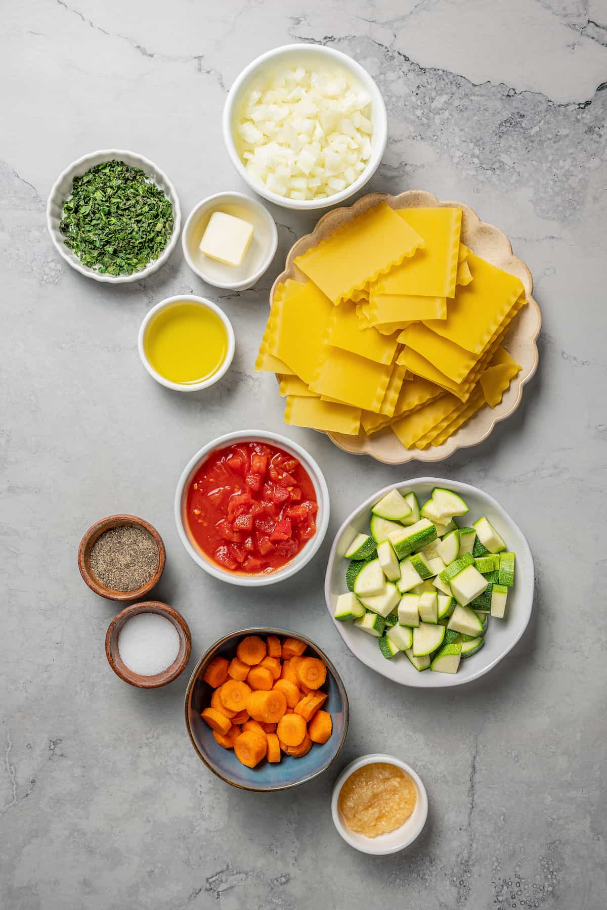 Overhead view of the ingredients needed for skillet lasagna: a plate of lasagna noodles, a bowl of zucchini, a bowl of carrots, a bowl of canned tomatoes, a bowl of onions, a bowl of basil, a bowl of garlic, a bowl of butter, a bowl of olive oil, a bowl of salt, and a bowl of pepper