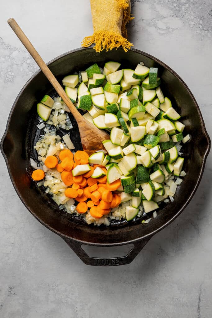 A skillet with raw zucchini and carrots on top of cooked onions and garlic, with a wooden spoon