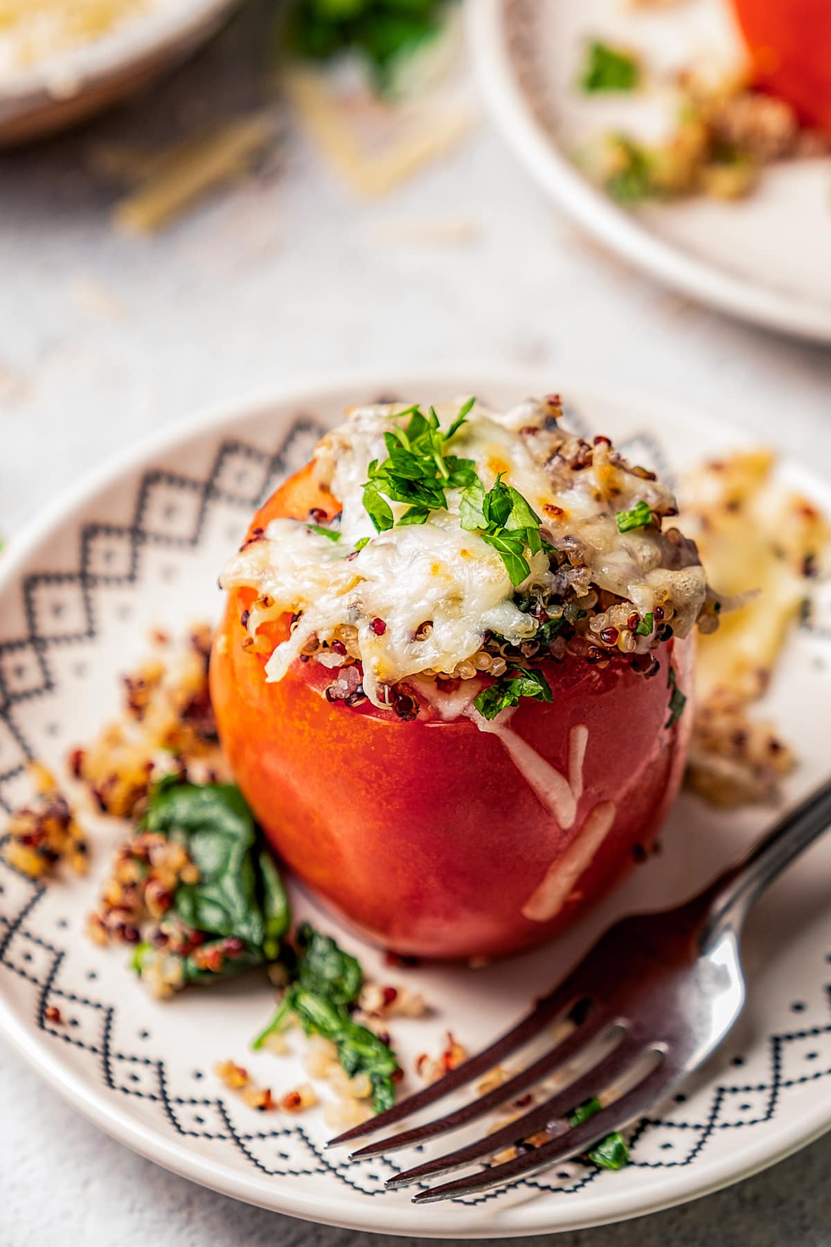 A quinoa stuffed tomato topped with melted cheese on a plate next to a fork, with more tomatoes served on plates in the background.