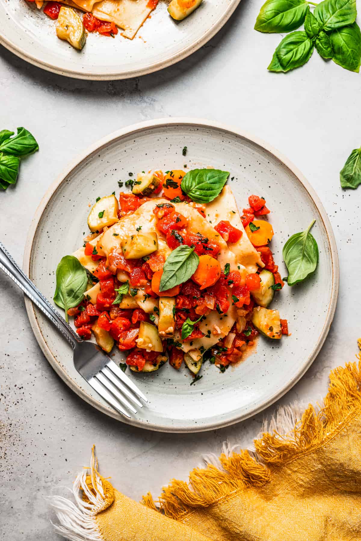 Overhead view of a plate of lasagna topped with fresh basil, and a fork placed on the dinner plate.