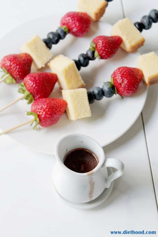 A small white pitcher filled with chocolate sauce next to fruit and cake on skewers 