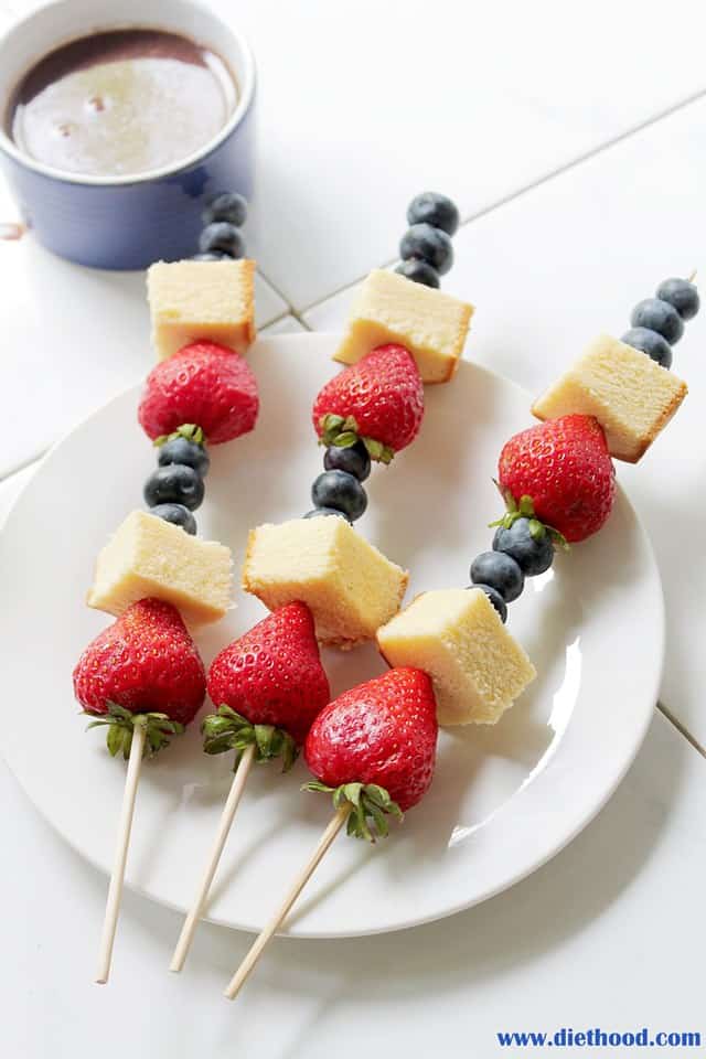 Strawberries, blueberries and pound cake on skewers on a white plate with chocolate sauce next to it