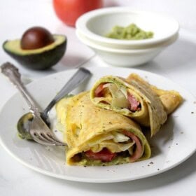 An egg omelette wrapped up with bacon, lettuce, tomato and avocado on a white plate