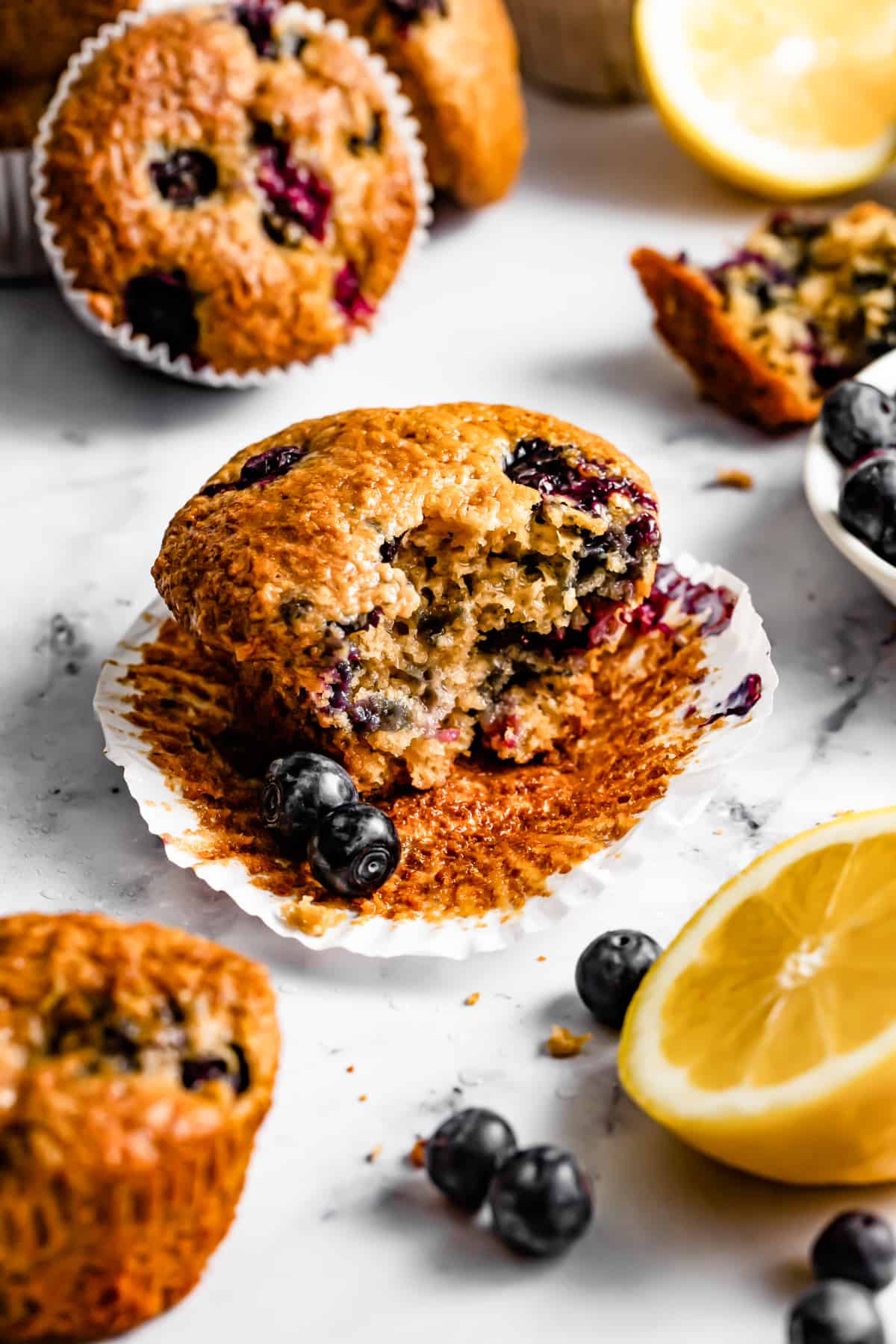 Blueberry lemon muffins surrounded by fresh blueberries. One muffin has a bite taken out of it.