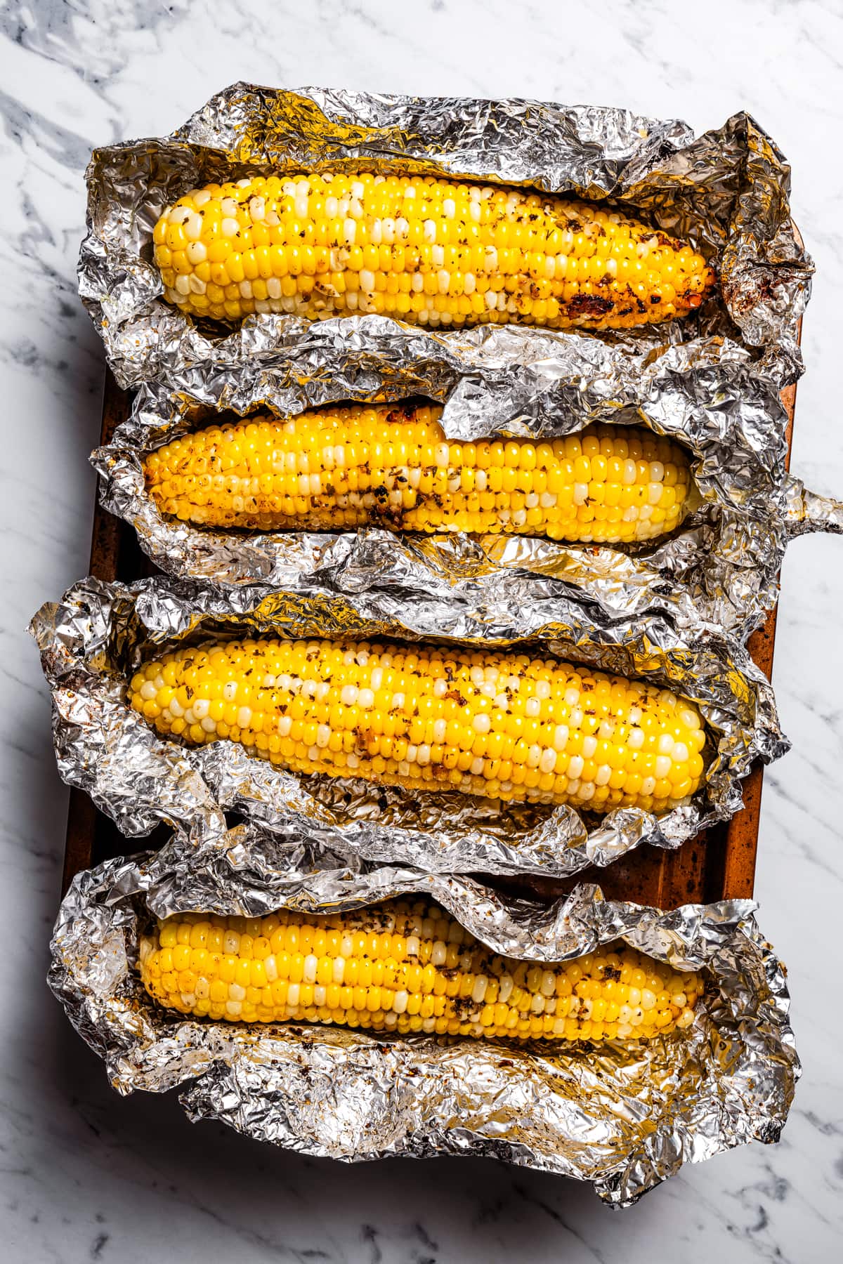 Four ears of corn partially unwrapped in foil on a baking sheet.