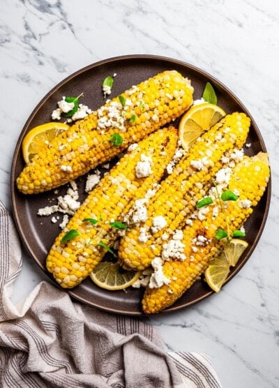 Overhead view of four ears of corn on a plate topped with goat cheese and garnished with lemon wedges.