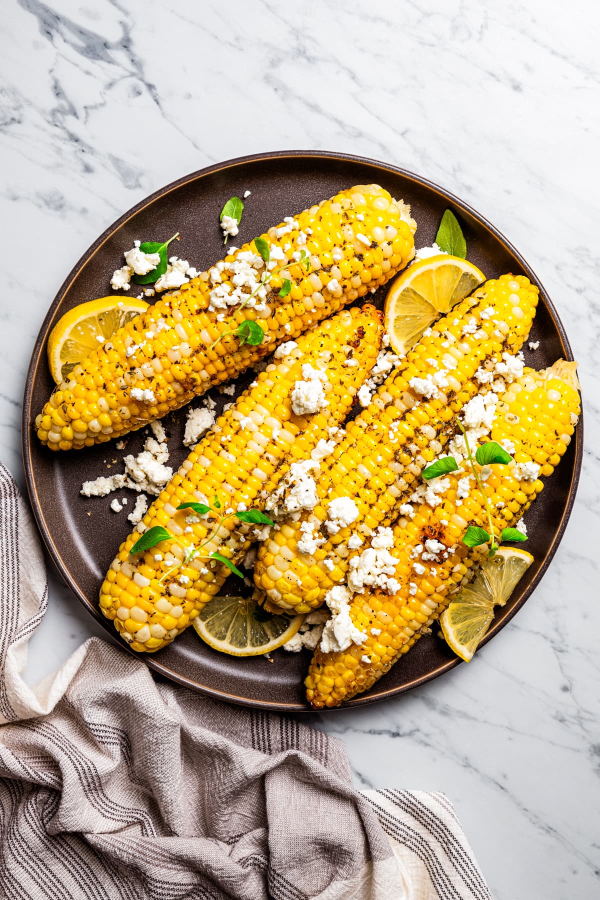 Overhead view of four ears of corn on a plate topped with goat cheese and garnished with lemon wedges.