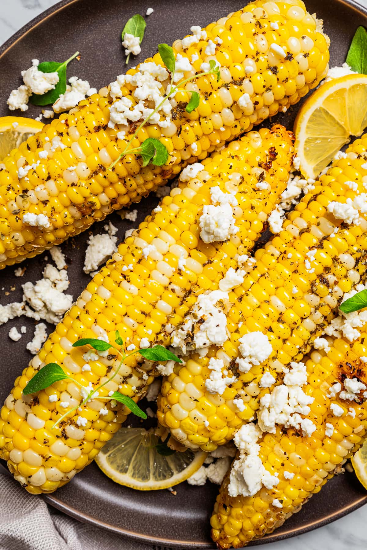 Close-up overhead view of grilled corn on the cob on a plate topped with goat cheese and garnished with lemon wedges.