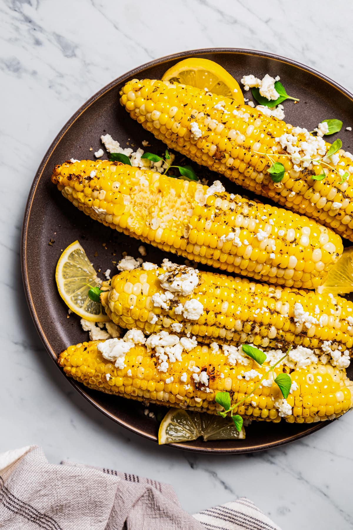 Overhead view of grilled corn on the cob on a plate topped with goat cheese and garnished with lemon wedges.