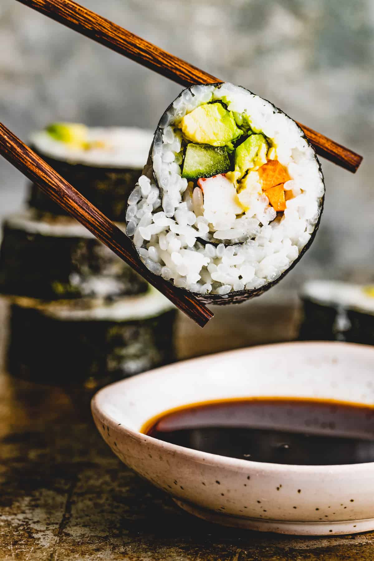 Chopsticks holding a slice of maki sushi over a bowl of soy sauce.