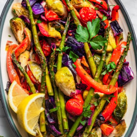 Roasted asparagus, bell peppers, tomatoes, cabbage, and brussel sprouts served on a platter with lemon wedges.