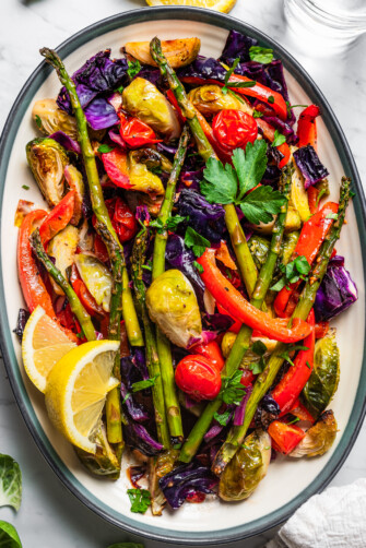 Roasted asparagus, bell peppers, tomatoes, cabbage, and brussel sprouts served on a platter with lemon wedges.