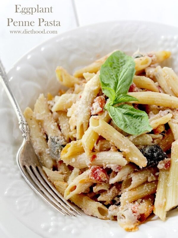 A close up of penne pasta in a white bowl with tomatoes, cheese, eggplant and topped with basil