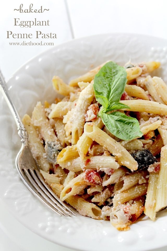 A close up of penne pasta in a white bowl with tomatoes, cheese, eggplant and topped with basil 