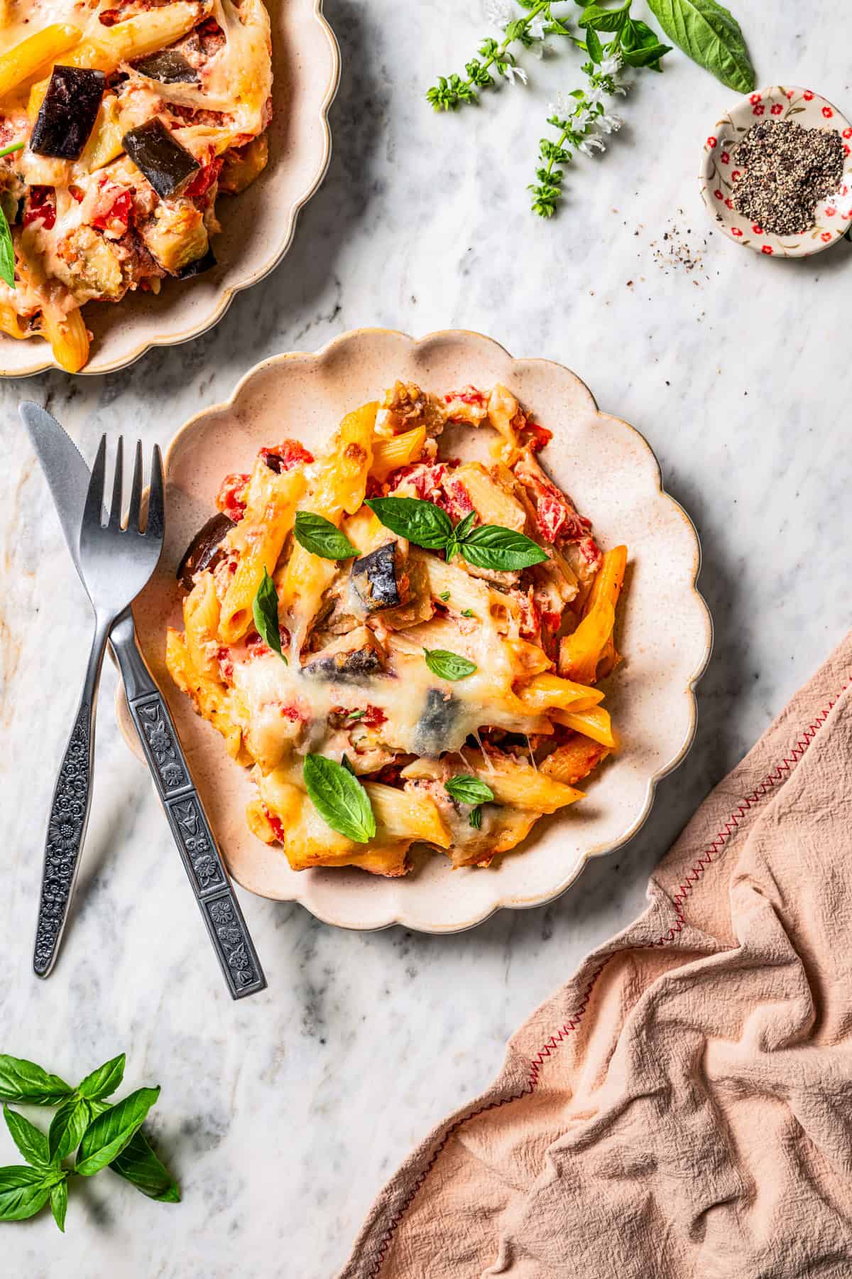 Two servings of baked penne pasta garnished with fresh basil and served on a dinner plate next to a fork and knife.