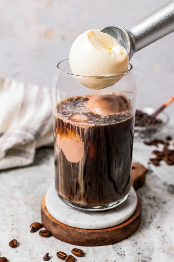 Adding a scoop of vanilla ice cream to black coffee in a drinking glass.