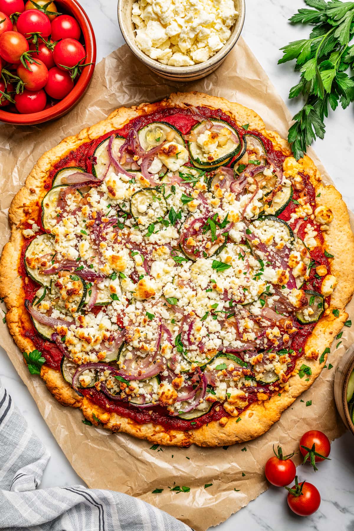 Overhead view of pizza topped with zucchini slices and goat cheese and surrounded by bowls filled with pizza toppings.