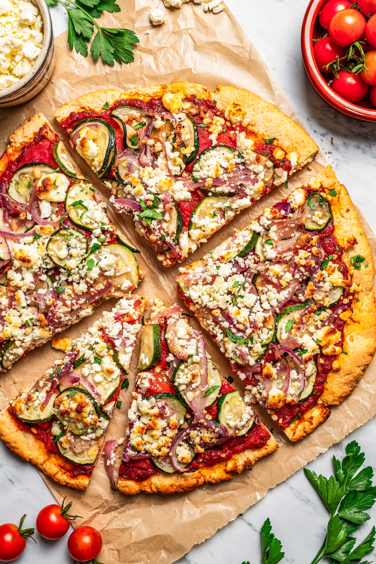 Overhead view of zucchini and goat cheese pizza cut into slices next to bowls of toppings.