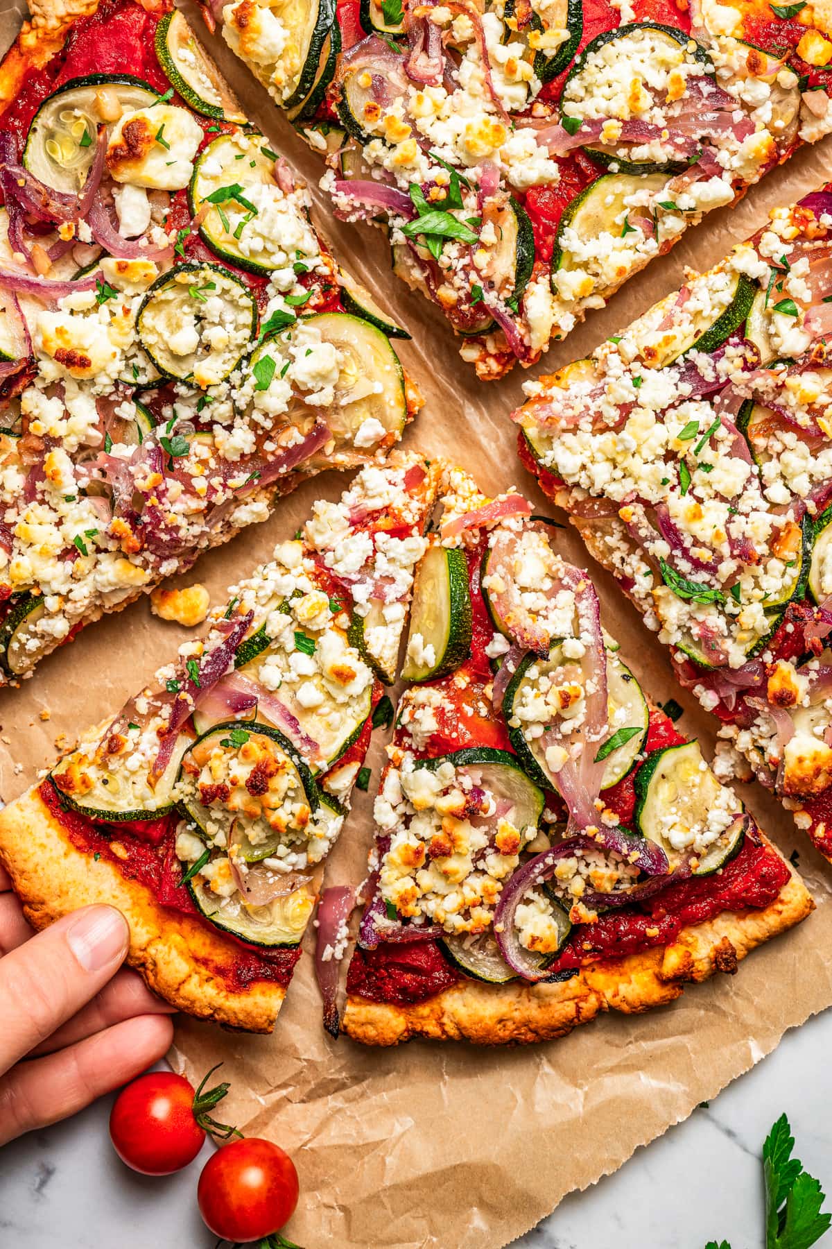 A hand lifts a slice from a zucchini goat cheese pizza.