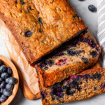 Overhead view of a loaf of blueberry banana bread cut into slices next to a bowl of blueberries.