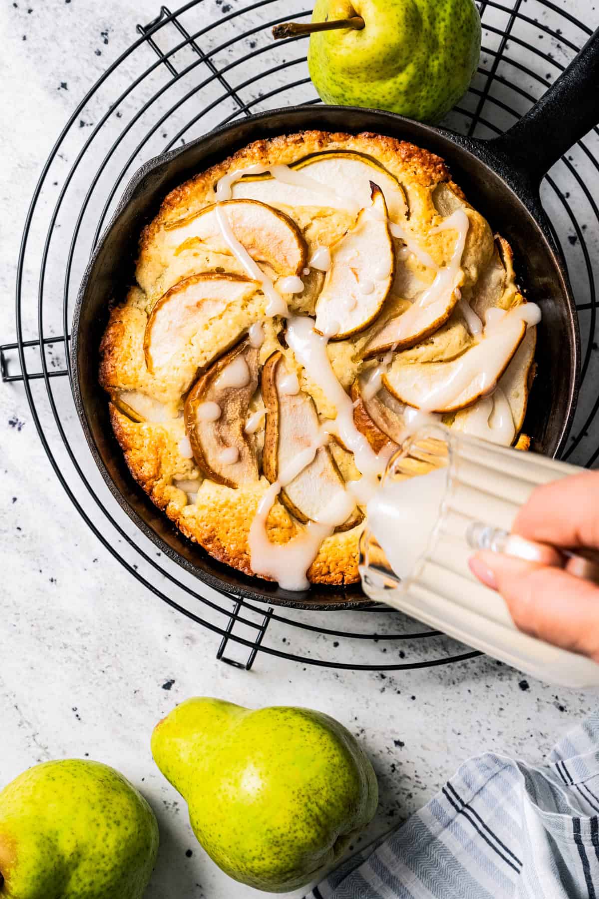 A hand pouring glaze over a freshly baked pear cake in a skillet, with a wire rack underneath and fresh pears on the side.