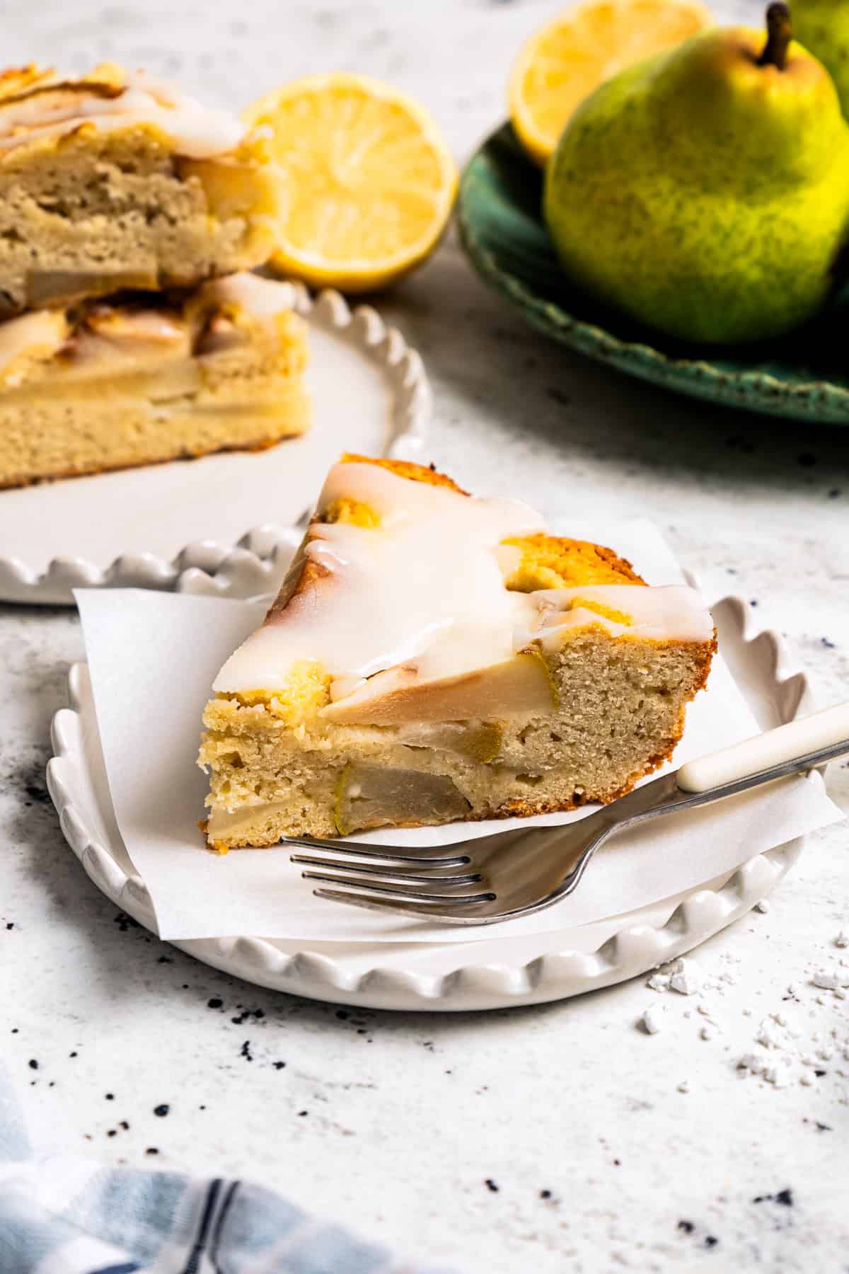 Side view of a slice of pear cake on a dessert plate, with pears placed in the background.