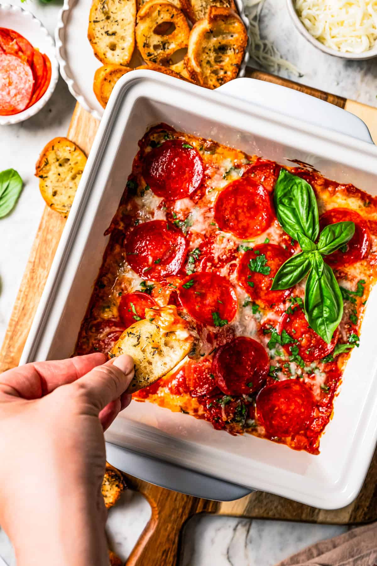 A hand scooping out cheesy pepperoni pizza dip from a baking dish.