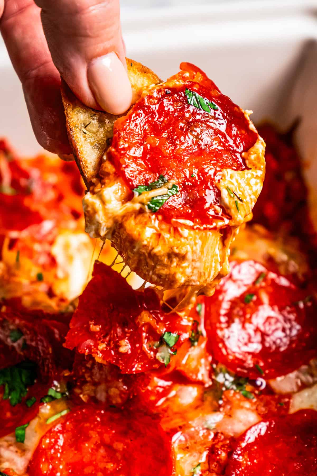 Close up of a hand holding up a slice of bread dipped in cheesy pepperoni pizza dip.