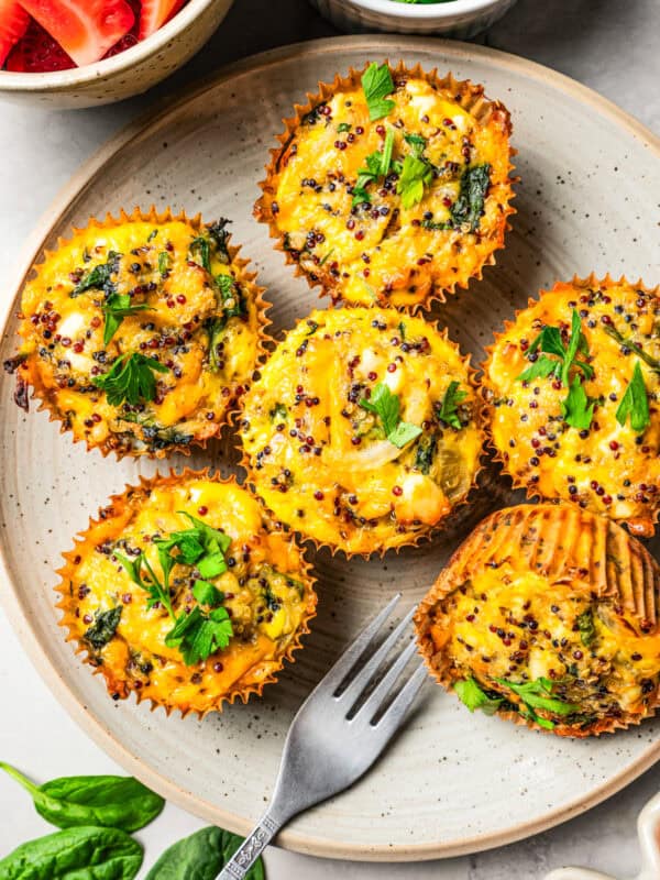 Six quiche muffins are arranged on a white plate, and a bowl of strawberries and fresh green herbs are set next to the plate.