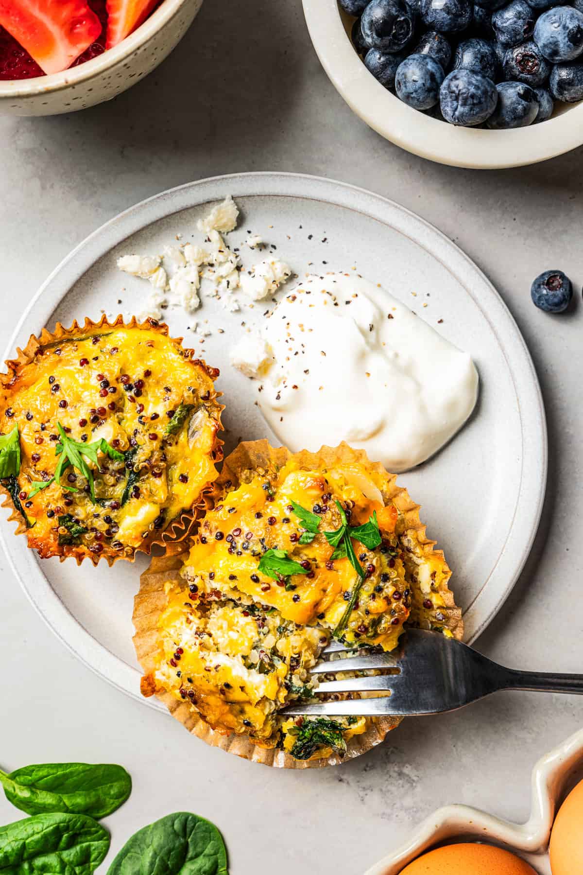 A fork is pulling apart a quiche muffin set next to a second muffin, a dollop of yogurt, and feta cheese on a plate, surrounded by bowls of berries.