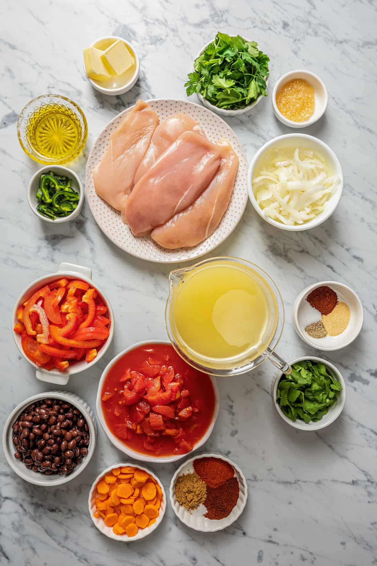 Overhead view of the ingredients needed for chicken tortilla soup: a bowl of butter, a bowl of parsley, a bowl of minced garlic, a plate of raw chicken breasts, a bowl of diced onion, a glass of olive oil, a bowl of sliced jalapeños, a bowl of sliced red bell peppers, a bowl of canned tomatoes, a bowl of canned black beans, a a bowl of carrots, two bowls of spices, a bowl of parsley, and a pyrex of chicken broth
