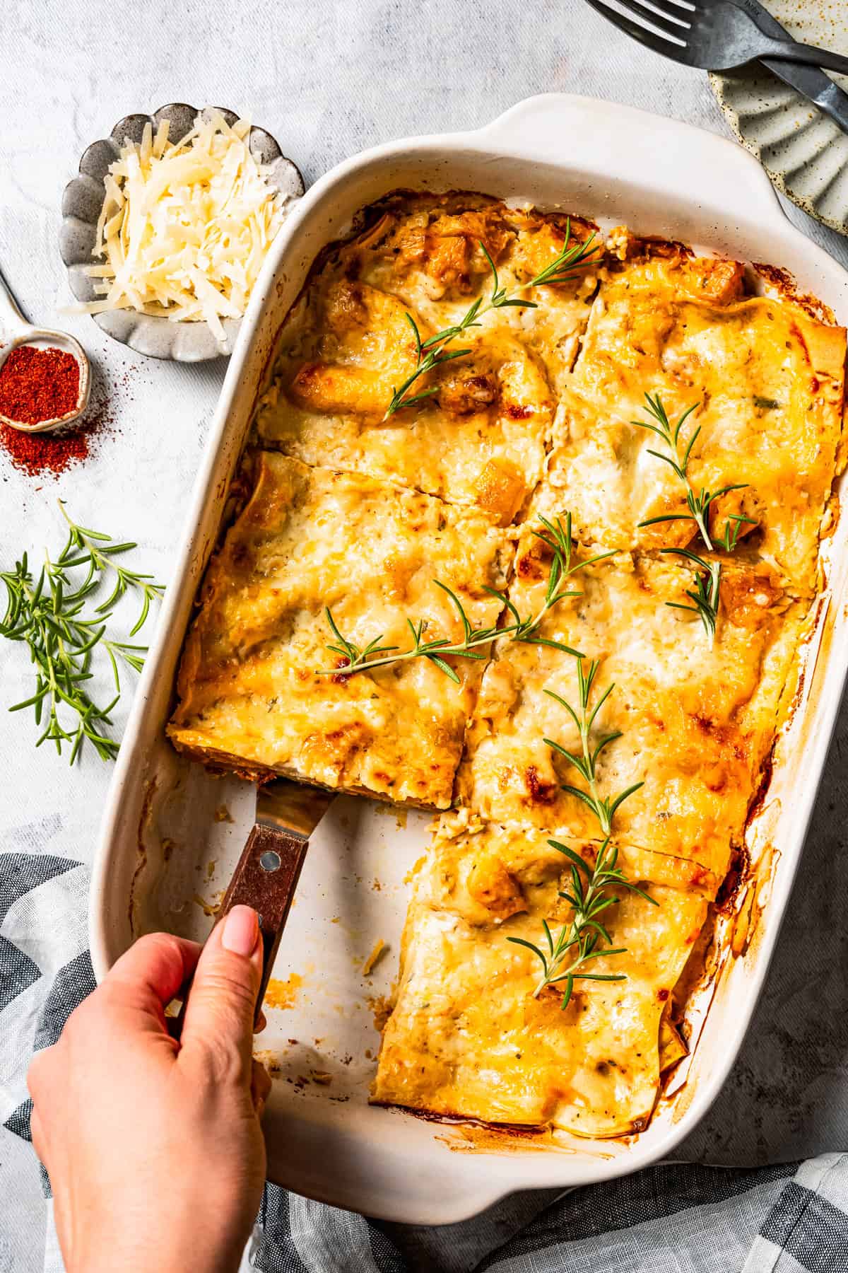 A hand sliding a spatula under a slice of butternut squash lasagna in a casserole dish.