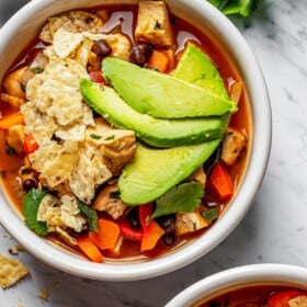 Overhead view of two bowls of tortilla soup topped with avocado and tortilla chips, next to a bowl of chips, a bowl of limes, half an avocado, and parsley.
