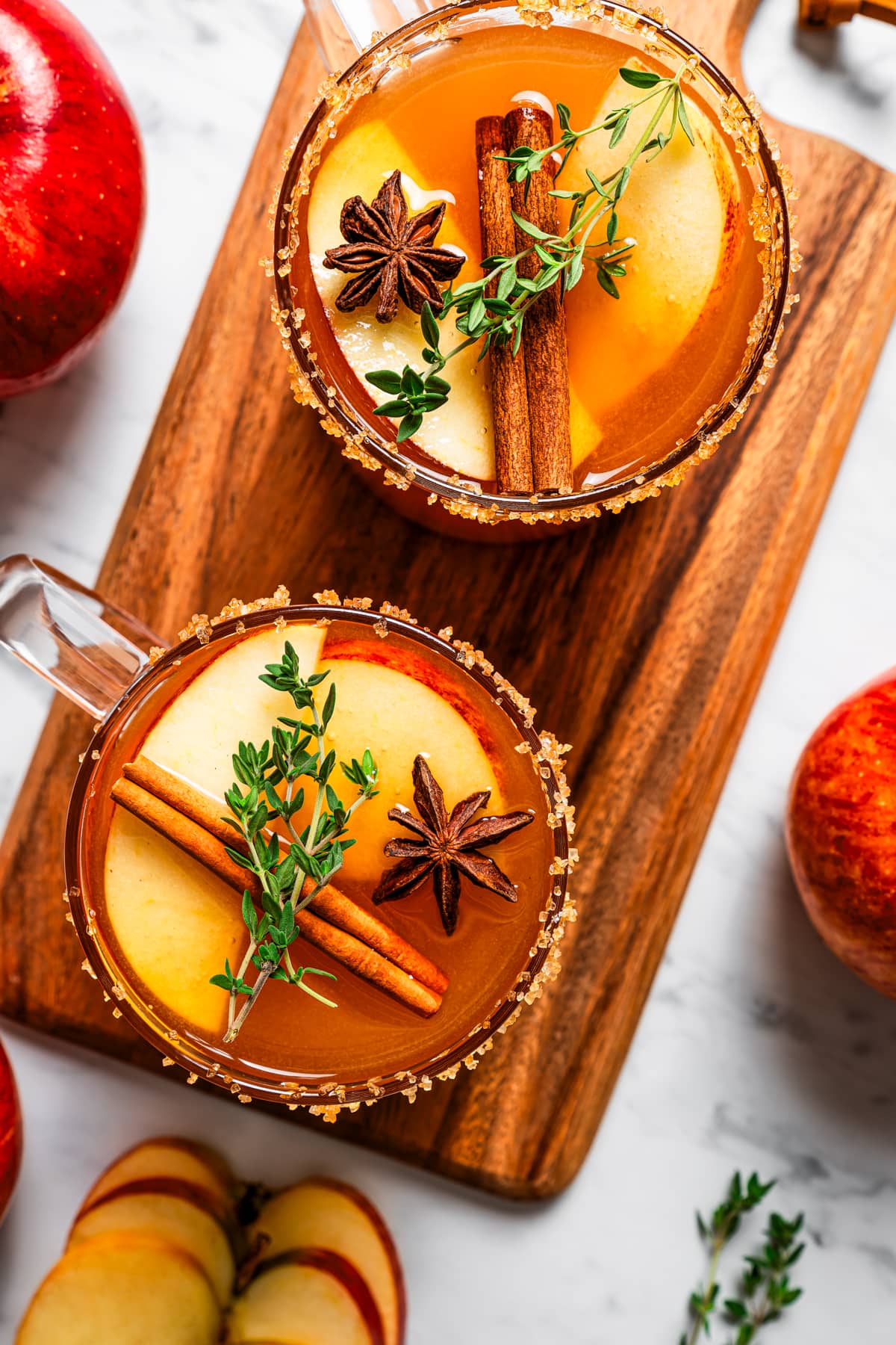 Overhead image of two glass mugs of spiked apple cider garnished with cinnamon sticks, apple slices, and greens.