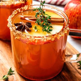 A glass cup filled with spiked apple cider garnished with apple slices, thyme, anise, and a cinnamon stick.