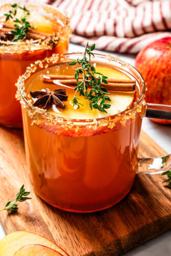 A glass cup filled with spiked apple cider garnished with apple slices, thyme, anise, and a cinnamon stick.