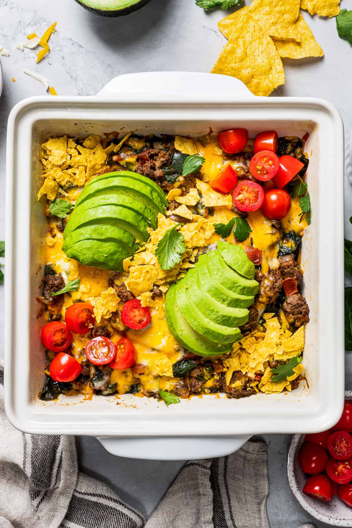Overhead view of a baked taco casserole in a square baking dish topped with sliced avocados, cherry tomatoes, and fresh cilantro.