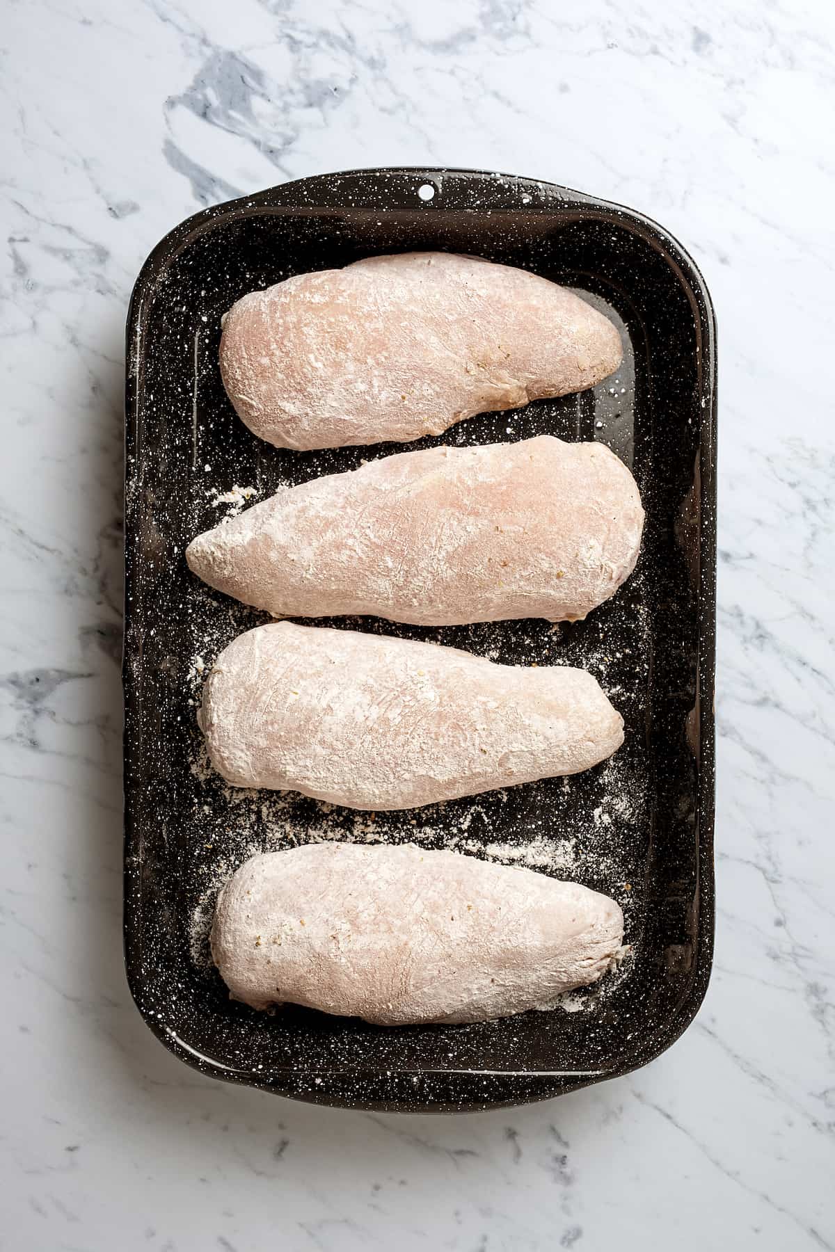 Four chicken breasts lightly coated in flour, lined up on a black baking dish.